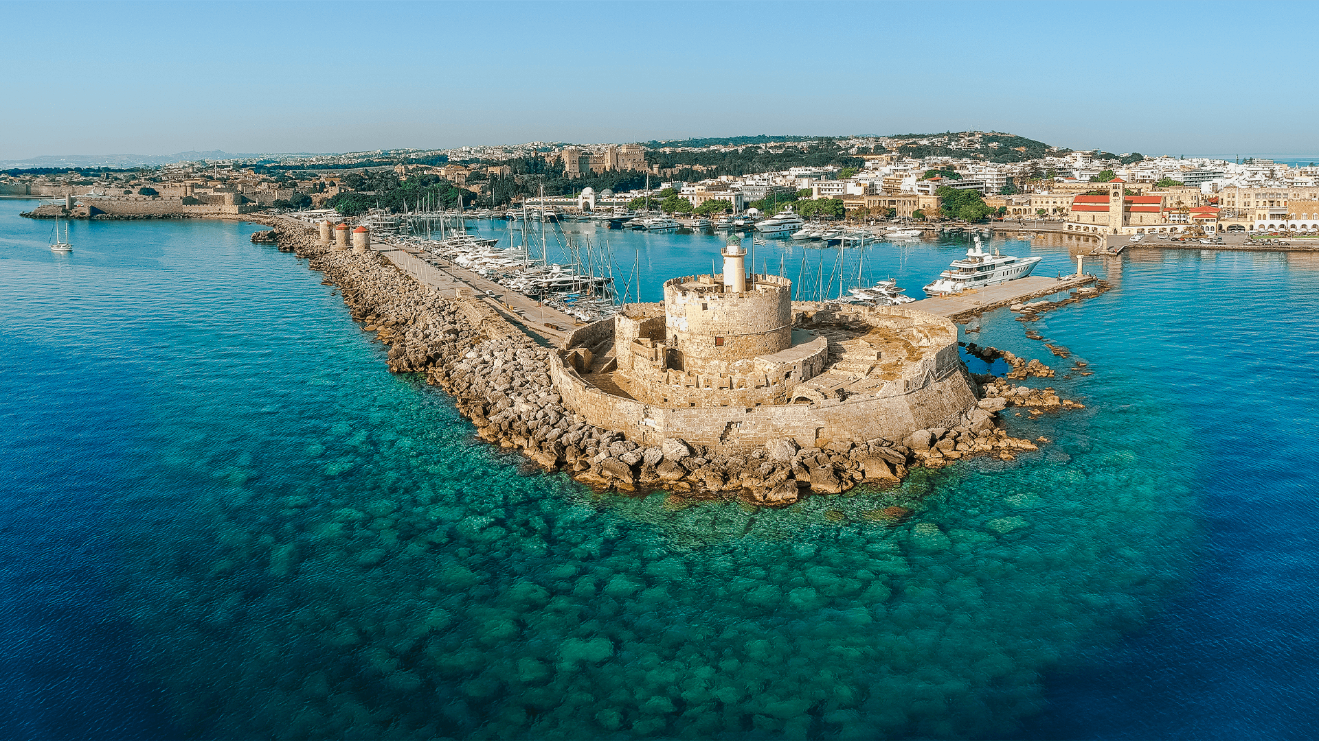 Mandraki Harbour in Rhodes, Greece, with boats moored along the waterfront and the historic city rising in the background.